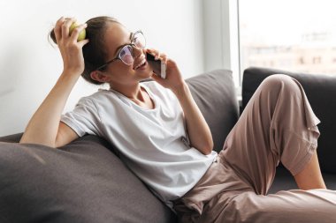 Gözlüklü, cep telefonuyla konuşan ve oturma odasında kanepede otururken elma tutan güzel bir kadın.