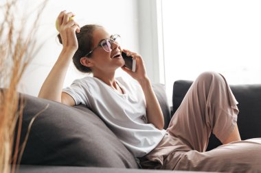 Gözlüklü, cep telefonuyla konuşan ve oturma odasında kanepede otururken elma tutan heyecanlı bir kadın resmi.