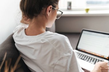 Gözlüklü esmer kadının oturma odasındaki kanepede otururken dizüstü bilgisayarda yazı yazması.