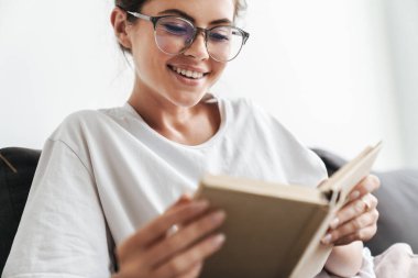 Gözlüklü neşeli beyaz kadının oturma odasındaki kanepede otururken kitap okuduğu bir resim.