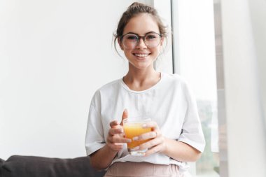 Gözlüklü, gülümseyen, kameraya bakan ve salonda meyve suyu içen bir kadın resmi.