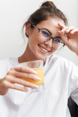 Gözlüklü güzel bir kadının oturma odasında kanepede otururken kameraya bakıp meyve suyu içmesi.