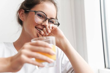 Gözlüklü, gülümseyen, kameraya bakıp salonda meyve suyu içen tatlı bir kadın resmi.