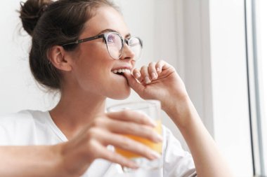 Gözlüklü, gülümseyen, hayal kuran ve salonda meyve suyu içen güzel bir kadın resmi.