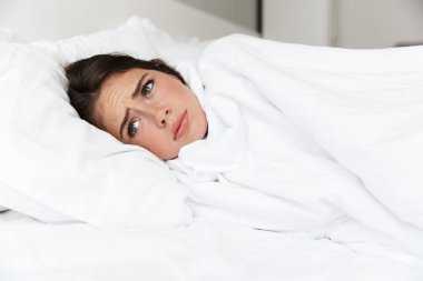 Üzgün, genç bir kız yatak odasında, battaniyeye sarılmış bir şekilde yatıyor.