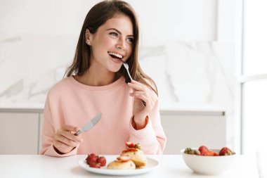 Mutfak masasında otururken güzel, gülümseyen, sağlıklı bir kahvaltı yapan genç kız.