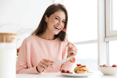 Mutfak masasında otururken güzel, gülümseyen, sağlıklı bir kahvaltı yapan genç kız.