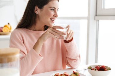Mutfak masasında oturmuş süt içerken güzel, güler yüzlü, sağlıklı bir kahvaltı yapan genç bir kızın fotoğrafını çek.