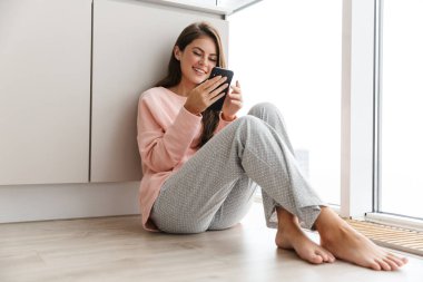 Mutfakta otururken cep telefonu kullanan çekici, gülümseyen bir genç kız.