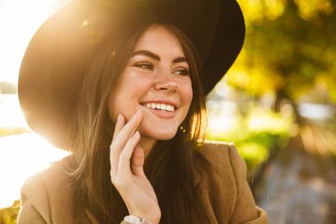 Sonbahar parkında bankta oturan ceketli ve şapkalı mutlu esmer kadının portresi.