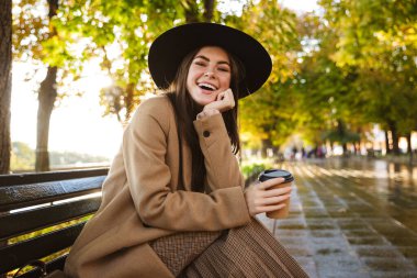 Paltolu ve şapkalı neşeli kadın portresi sonbahar parkında kahve fincanıyla bankta oturuyor.