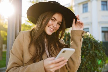 Açık havada yürürken elinde akıllı telefon tutan sonbahar ceketli ve şapkalı baştan çıkarıcı kadının portresi.
