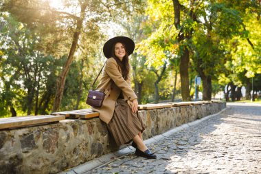 Sonbahar ceketli ve şapkalı esmer bir kadın yeşil parkta yürürken bankta oturuyor.