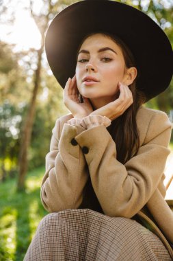 Yeşil parktaki bankta oturan sonbahar ceketli ve şapkalı baştan çıkarıcı kadın.