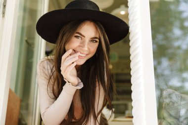 Kafenin penceresinden dışarı bakarken gülümseyen şapkalı genç bir kadın görüntüsü