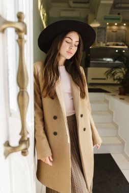 Güz ceketli ve şapkalı genç bir kadının kafenin açık kapısının önünde duruşu.