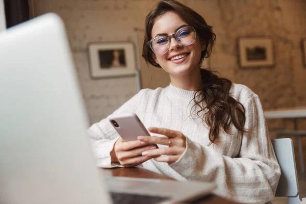 カフェでノートパソコンやスマートフォンを使って眼鏡をかける若い美人白人女性の姿 — ストック写真
