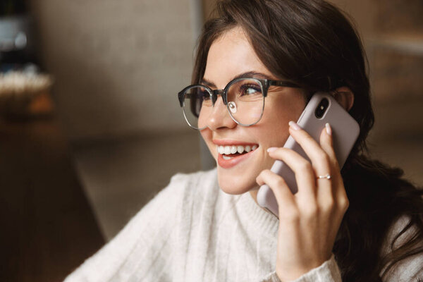 Image of young beautiful caucasian woman wearing eyeglasses talking on smartphone in cafe