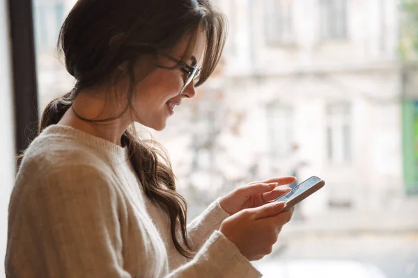 スマートフォンで眼鏡をかけ カフェの窓のそばに立つ若い美しい白人女性のイメージ — ストック写真