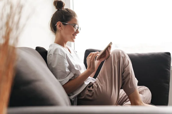 Image of beautiful woman in eyeglasses smiling and typing on cellphone while sitting on sofa at living room