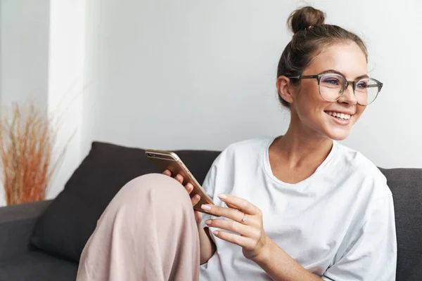 Image of young woman in eyeglasses smiling and using cellphone while sitting on sofa at living room