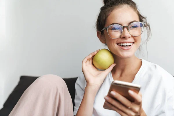 リビングでソファに座りながら 携帯電話を使って眼鏡をかけ リンゴを持っている素敵な女性の笑顔のイメージ — ストック写真