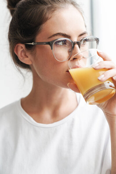 Image of young nice woman in eyeglasses looking at camera and drinking juice at living room