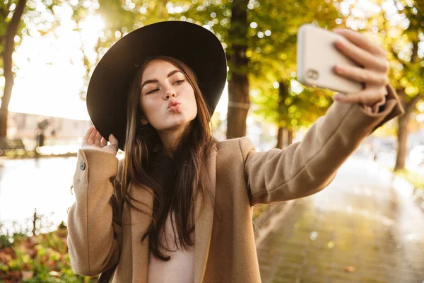 Portrait of seductive woman in coat and hat taking selfie photo on cellphone while walking in autumn park