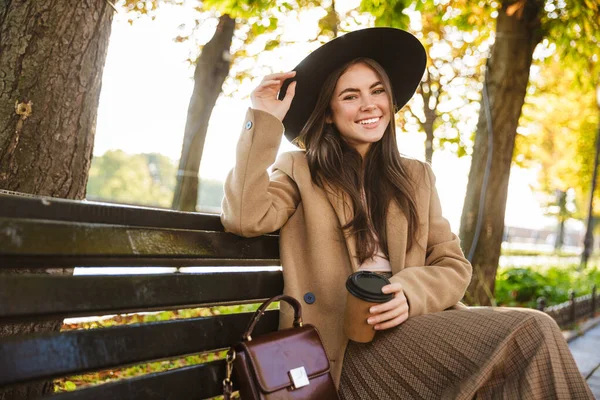 秋天公园里穿着外套 头戴帽子 头戴咖啡杯坐在长椅上的雅致女子的画像 — 图库照片