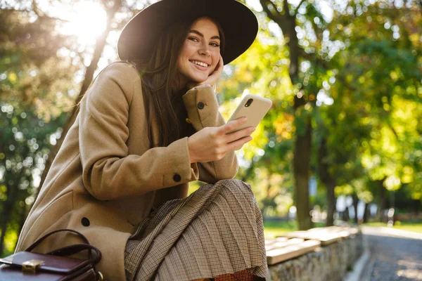 Obrázek Pozitivní Ženy Podzimním Kabátu Klobouku Pomocí Smartphonu Při Sezení — Stock fotografie