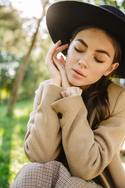 秋のコートと帽子の魅力的な女性のイメージは屋外の緑の公園のベンチに座って — ストック写真