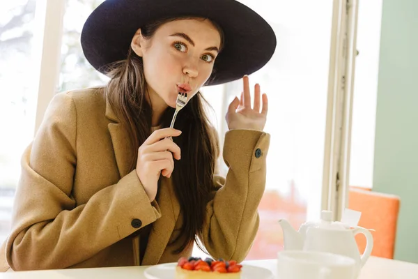 身着秋衣头戴礼帽的年轻女子在室内咖啡馆吃甜蛋糕的照片 — 图库照片