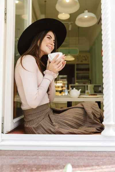 Imagen Mujer Complacida Sombrero Sonriendo Mientras Bebe Taza Sentado Alféizar — Foto de Stock