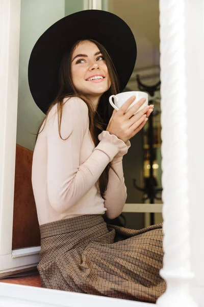 Imagen Mujer Caucásica Sombrero Sonriendo Mientras Bebe Taza Sentado Alféizar —  Fotos de Stock