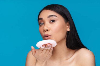 Asyalı genç ve güzel bir kadının mavi duvar arkasında poz verirken çekilmiş fotoğrafı. Elinde pudra ponponu var..