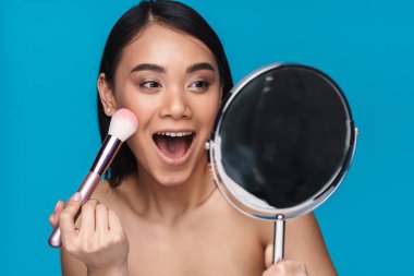 Asyalı iyimser genç bir kadının mavi duvar arkasında poz verirken çekilmiş fotoğrafı. Elinde ayna tutan fırça tozu uygula..