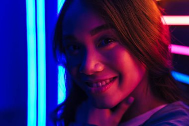 Asyalı mutlu, mutlu, gülümseyen, güzel, neon ışıkların üzerinde poz veren genç bir kadın..