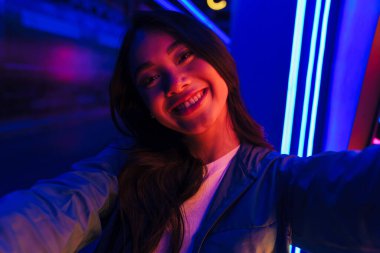 Neon ışıklarının üzerinde poz veren neşeli genç bir kadının fotoğrafı. Kamerayla selfie çek..
