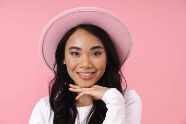 Şapka takan, gülümseyen ve pembe arka planda izole edilmiş kameraya bakan Asyalı genç esmer kadın.