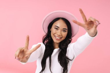 Şapka takan, gülümseyen ve pembe arka planda izole edilmiş barış parmaklarını gösteren Asyalı genç esmer kadın.