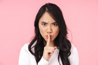 Kaşlarını çatan ve pembe arka planda izole edilmiş dudaklarda parmak tutan Asyalı genç esmer kadın.