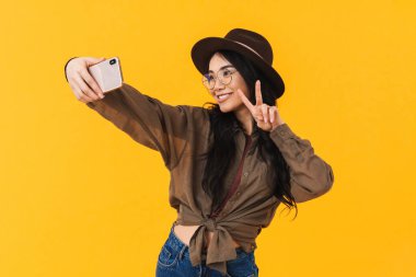Sarı arka planda izole edilmiş cep telefonuyla fotoğraf çeken Asyalı genç esmer kadın.