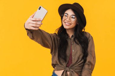 Sarı arka planda izole edilmiş cep telefonuyla fotoğraf çeken Asyalı genç esmer kadın.