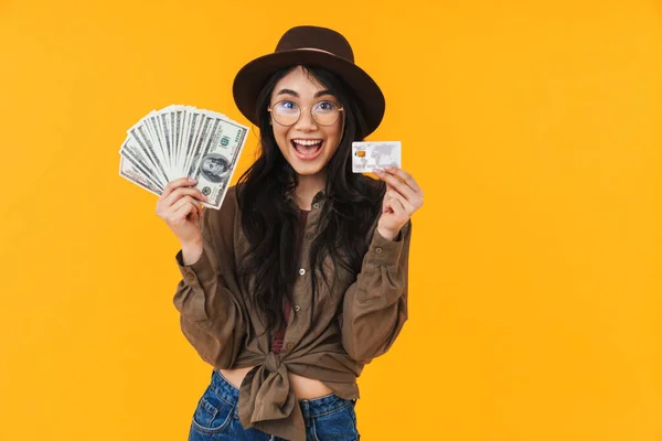 照片上年轻的黑发亚洲女人头戴一顶帽子 手里拿着钞票和信用卡 背景呈黄色 — 图库照片