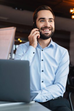 Beyaz gömlekli yakışıklı iş adamının portresi cep telefonuyla konuşuyor ve ofiste çalışırken dizüstü bilgisayar kullanıyor.