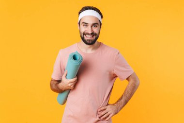 Tişörtlü, kaslı, atletik, gülümseyen ve sarı arka planda izole edilmiş spor minderi taşıyan genç bir adam resmi.