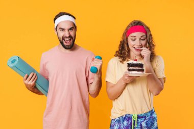 Sağlıksız bir pasta tutan kadın sarı arka planda izole haldeyken spor ayakkabısı ve dambıl tutan genç adamın portresi.