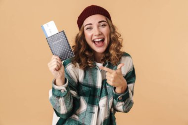 Örgü şapkalı ve elinde bej arka planda izole edilmiş pasaportu işaret eden güzel, heyecanlı bir kadının fotoğrafı.