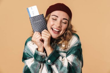 Örgü şapkalı, elinde biletleri olan ve bej arka planda sırıtan mutlu bir kadının fotoğrafı.