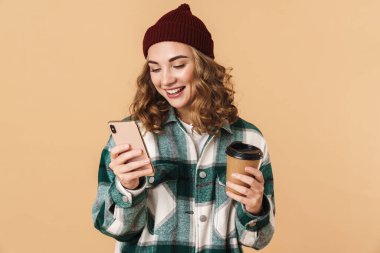 Örgü şapkalı neşeli bir kadın cep telefonu kullanıyor ve bej arka planda izole edilmiş kahve içiyor.